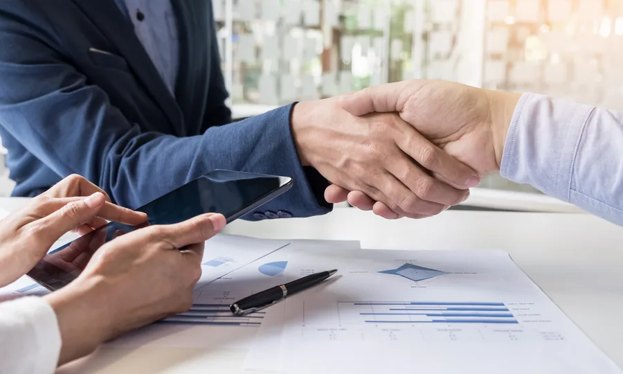 Two people shaking hands across a desk with printed reports and a tablet