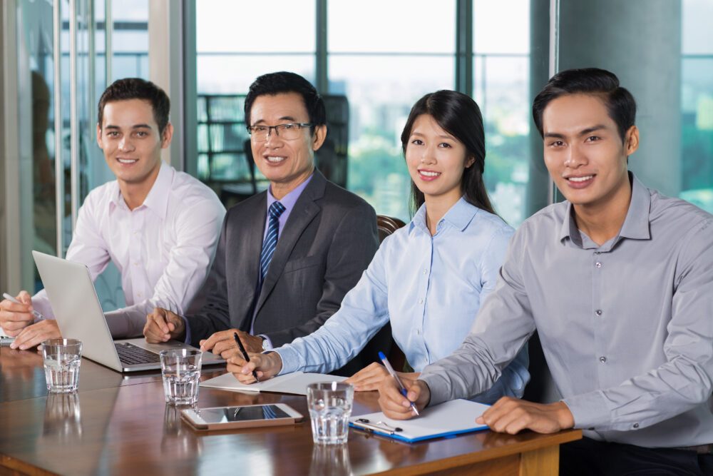 Four business professionals seated at a meeting table with notebooks, a laptop and glasses of water