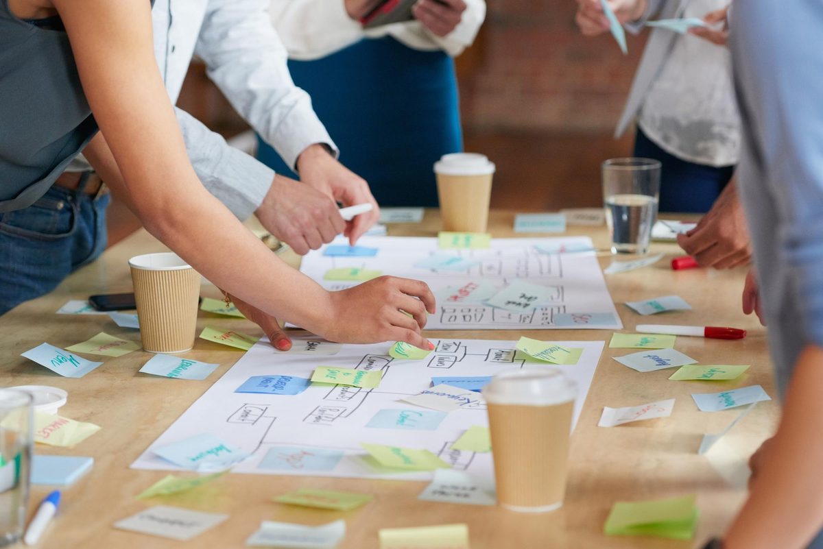 Colleagues arranging sticky notes on a hand-drawn planning map, with coffee cups and markers on the table